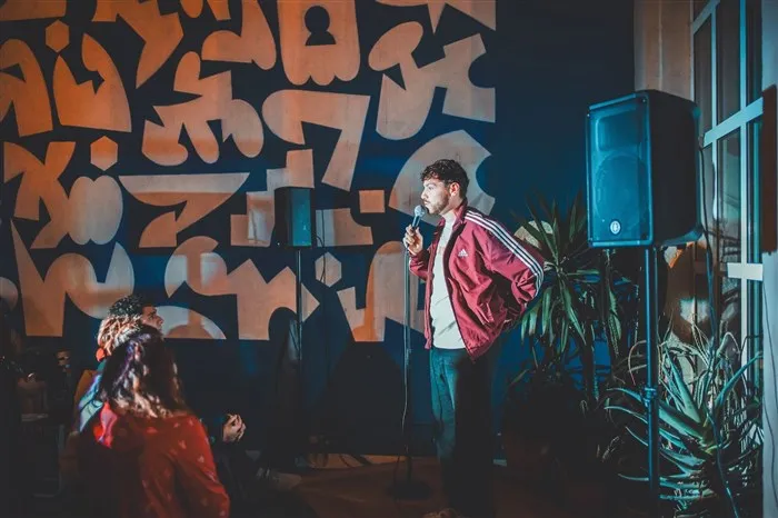 Un humoriste au micro sur la scène du Sunny Comedy Club, devant le mur graffé du The People